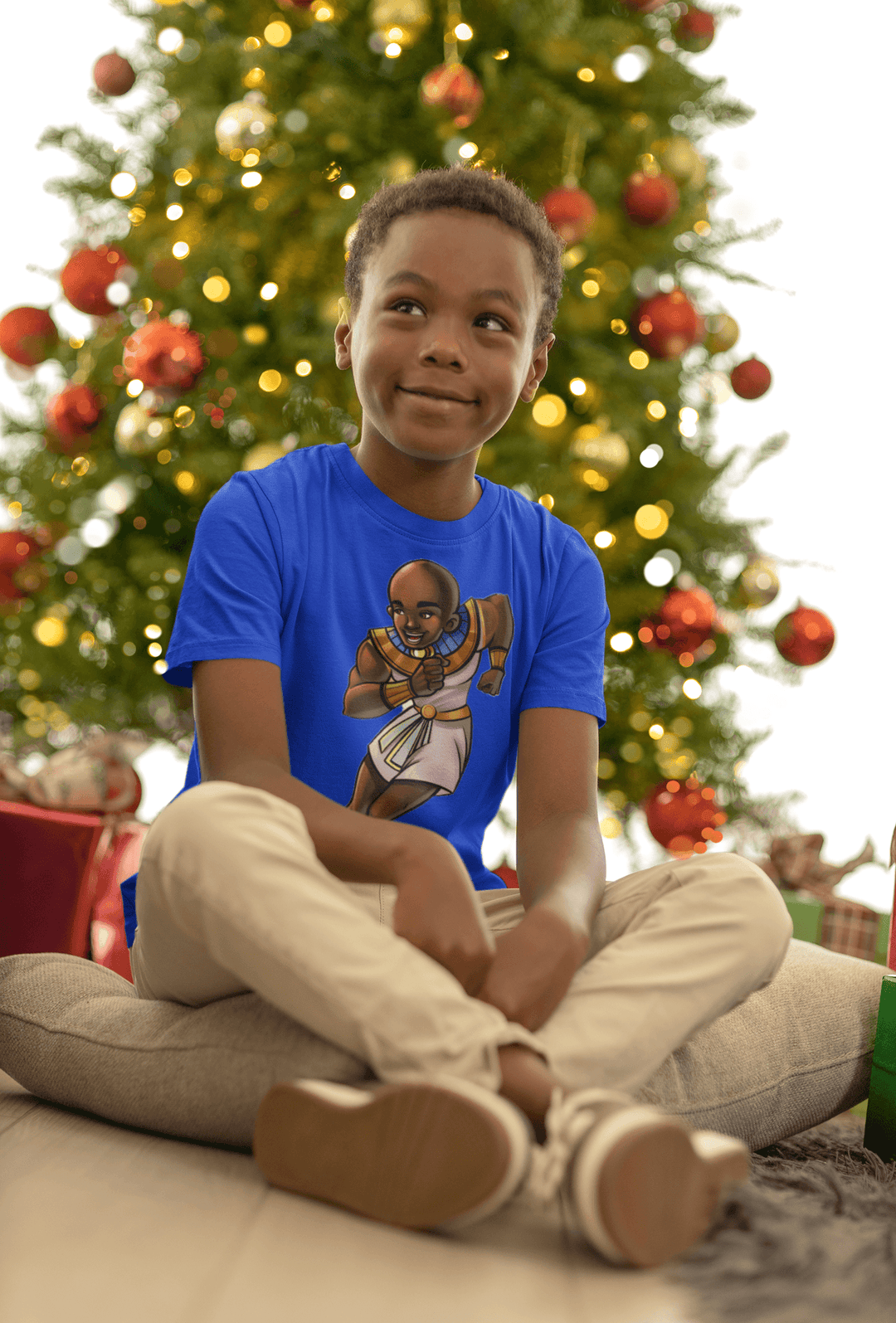 Boy wearing Ramses T-shirt from TL Johnson’s Mini Monarchs collection celebrating intelligence and royal leadership, sitting under a Christmas tree.