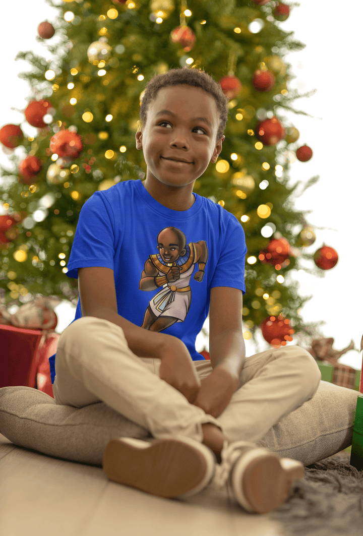 Boy wearing Ramses T-shirt from TL Johnson’s Mini Monarchs collection celebrating intelligence and royal leadership, sitting under a Christmas tree.
