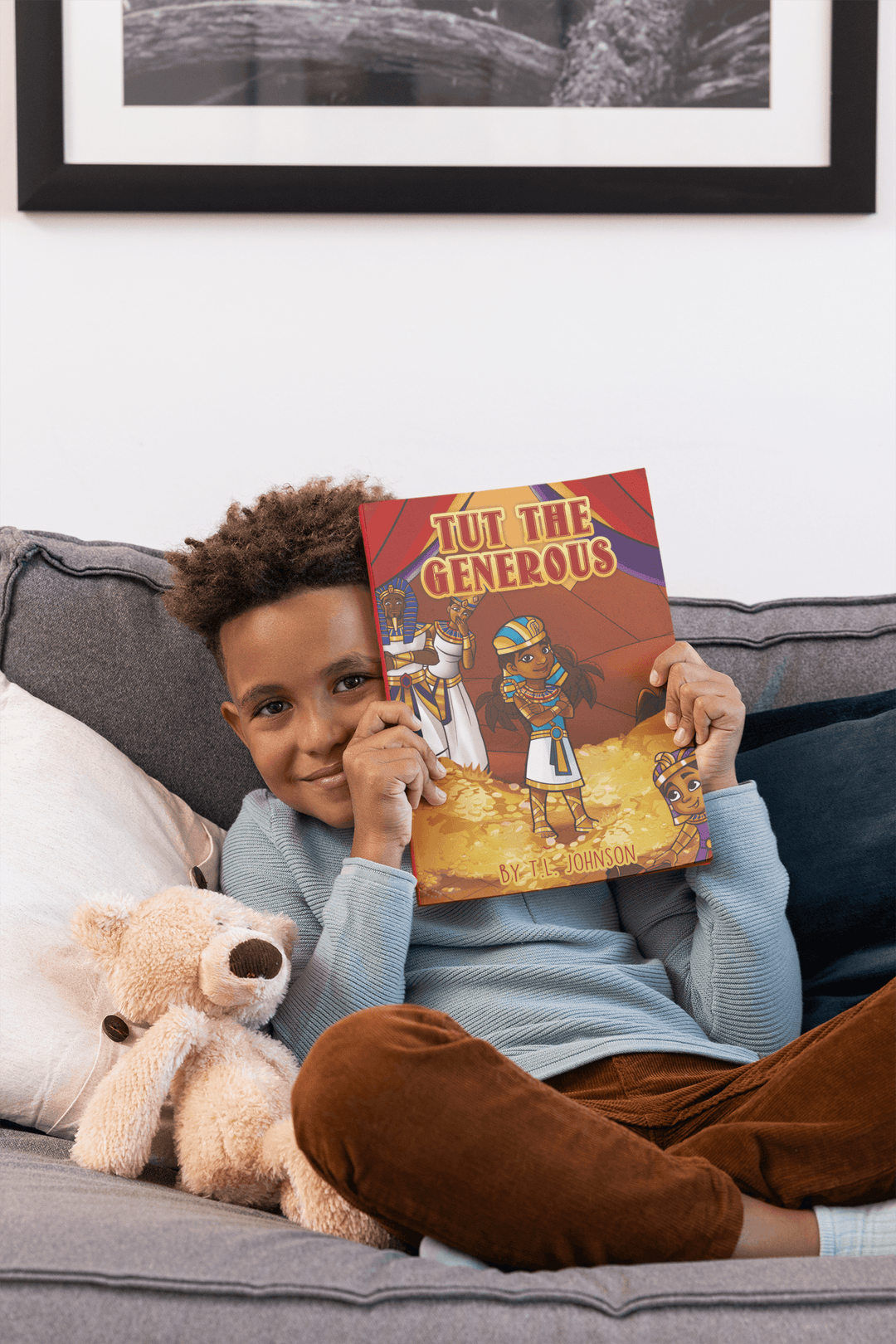 Young black boy smiling while holding Tut The Generous. on a couch next to his teddy bear